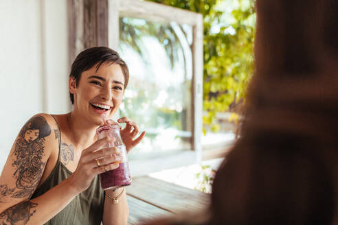 Frau mit Smoothie-Glas im Gespräch mit ihrer Freundin in einem Restaurant. Frau mit Tattoo auf der Hand in einem Restaurant, die Saft trinkt. - JLPSF00827