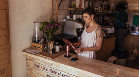 Woman standing at the billing counter of her coffee shop. Coffee shop owner operating the billing machine while a worker prepares coffee in the background. - JLPSF00830