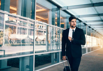 Young businessman on business trip walking with luggage and looking away. Business executive carrying suitcase in airport. - JLPSF00850