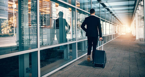 Rear view of young businessman walking outside public transport building with luggage. Business traveler pulling suitcase in modern airport terminal. - JLPSF00852