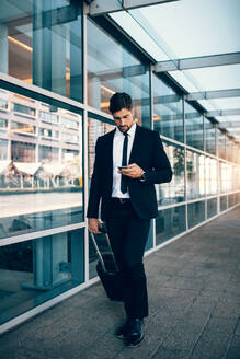 Gutaussehender Geschäftsmann, der ein Mobiltelefon benutzt, während er mit einem Koffer am Flughafen spazieren geht. Junger Mann, der ein Mobiltelefon mit Gepäck im Flughafenterminal benutzt. - JLPSF00854