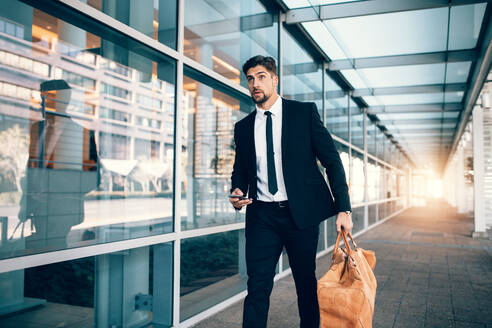 Gutaussehender Geschäftsreisender, der mit einer Handtasche und einem Handy in der Hand am Flughafen-Terminal spazieren geht. Kaukasischer Geschäftsmann, der eine Tasche mit einem Smartphone am Flughafen trägt. - JLPSF00856