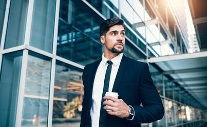 Businessman walking outside public transport building with a coffee and looking away. Handsome young man on a business trip. - JLPSF00857