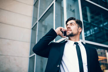 Portrait of handsome young man talking on mobile phone and looking away. Caucasian businessman making a phone call outside a building. - JLPSF00861