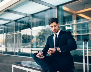 Junger Geschäftsmann wartet und prüft die Zeit mit seinem Handy. Geschäftsreisender sitzt auf einer Bank und schaut auf seine Uhr im Flughafenterminal. - JLPSF00863