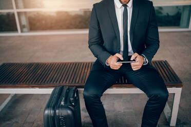 Ausschnitt eines jungen Geschäftsmannes, der mit einem Koffer auf einer Bank sitzt und ein digitales Tablet benutzt. Ein Mann wartet in einer öffentlichen Verkehrsstation. - JLPSF00866