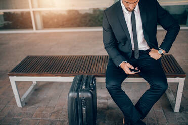 Junger Geschäftsmann, der mit seinem Koffer auf einer Bank sitzt und ein Smartphone benutzt. Geschäftsmann, der in einem Bahnhof auf öffentliche Verkehrsmittel wartet. - JLPSF00867