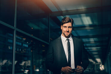Portrait of handsome young businessman looking at camera and smiling. He is standing in a modern building. - JLPSF00872