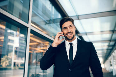 Geschäftsmann auf Geschäftsreise, der am Flughafen mit seinem Handy telefoniert. Kaukasischer Geschäftsmann, der ein Handy benutzt und lächelt. - JLPSF00876