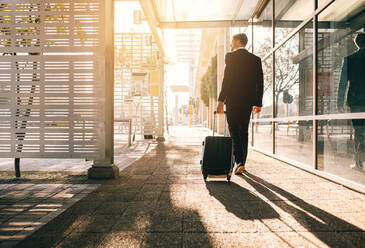 Rear view of businessman walking with bag outside airport. Young business traveler pulling suitcase on city street. - JLPSF00881