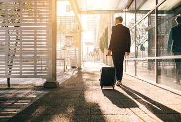 Rückansicht eines Geschäftsmannes, der mit einer Tasche außerhalb des Flughafens geht. Junger Geschäftsreisender, der einen Koffer auf einer Straße in der Stadt zieht. - JLPSF00881