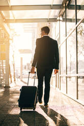 Junger Geschäftsmann mit Koffer vor dem Flughafengebäude. Vertikale Rückansicht eines männlichen Geschäftsreisenden, der seine Tasche auf einer Straße in der Stadt zieht. - JLPSF00882
