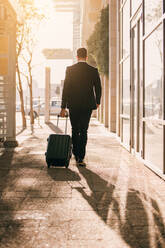 Vertical shot from behind of man walking with bag outside on city street. Young business traveler with suitcase outside airport. - JLPSF00883