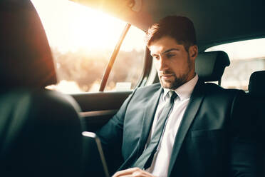 Gut aussehender Geschäftsmann, der einen Laptop benutzt, während er auf dem Rücksitz eines Autos sitzt. Kaukasischer männlicher Geschäftsmann, der in einem Auto reist und an einem Laptop arbeitet. - JLPSF00888
