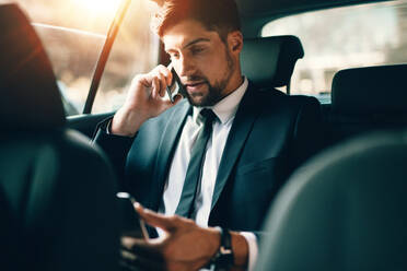 Junger Geschäftsmann, der mit einem Mobiltelefon spricht und einen Tablet-PC benutzt, während er auf dem Rücksitz eines Autos sitzt. Kaukasischer männlicher Geschäftsmann, der mit einem Taxi reist und auf ein digitales Tablet schaut. - JLPSF00889