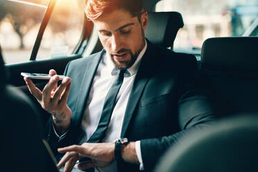 Handsome businessman talking on phone while sitting on the backseat of the car. Young man travelling by car using smart phone and checking time. - JLPSF00890