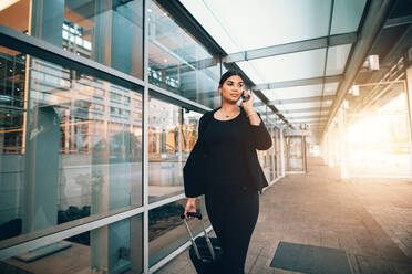 Frau auf Geschäftsreise, die mit ihrem Gepäck vor dem Flughafen spazieren geht und mit dem Handy telefoniert. Eine weibliche Reisende telefoniert. - JLPSF00896