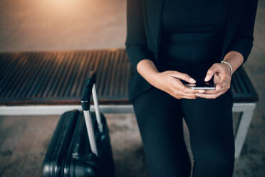 Ausschnitt einer jungen Geschäftsfrau, die mit einem Koffer auf einer Bank sitzt und ein Mobiltelefon benutzt, während sie in einer öffentlichen Verkehrsstation wartet. - JLPSF00897