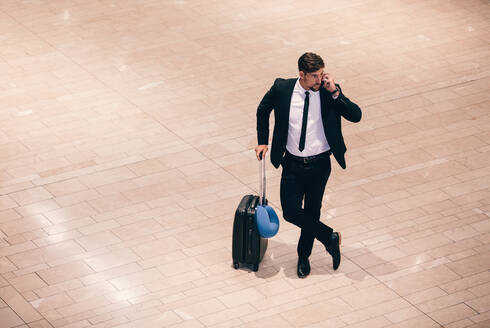 Top view shot of young businessman standing at airport terminal with suitcase and talking on phone. Geschäftsreisender beim Telefonieren während er auf seinen Flug wartet. - JLPSF00919