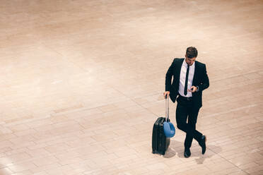 Top view shot of young businessman standing at airport terminal with suitcase and using mobile phone. Business traveler reading text message cell phone while waiting for his flight. - JLPSF00920