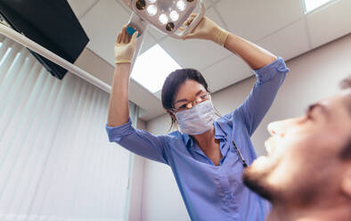 Dentist adjusting electric light while patient sitting on chair at dental clinic. Female doctor preparing for a dental treatment on male patient. - JLPSF00927