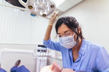 Zahnärztin mit Maske, die das Licht mit dem Patienten auf dem Stuhl in der Zahnklinik einstellt. Ärztin bereitet sich auf eine Zahnbehandlung bei einem männlichen Patienten vor. - JLPSF00929