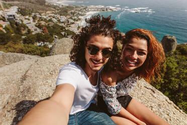 Young man and woman sitting together on top of cliff taking self portrait. Happy couple sitting on the top of mountain and taking selfie. - JLPSF00940