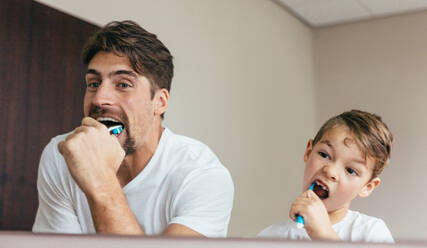 Father and son brushing teeth in bathroom. Young man with his son together brushing teeth and looking in mirror. - JLPSF00944