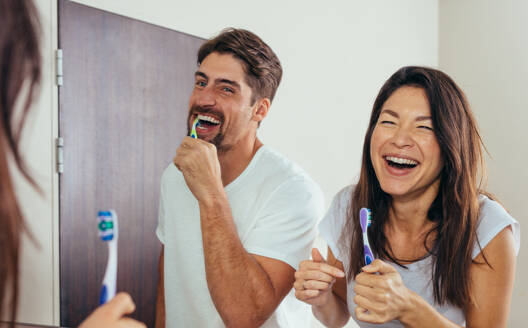 Lächelnder Mann und lächelnde Frau beim Zähneputzen im Badezimmer. Paar beim Zähneputzen vor dem Badezimmerspiegel und lachend. - JLPSF00950