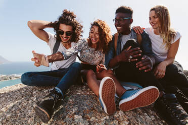 Eine Gruppe von Freunden sitzt zusammen auf einem Berggipfel und macht ein Selbstporträt mit dem Handy. Junge Männer und Frauen machen ein Selfie im Urlaub. - JLPSF00951