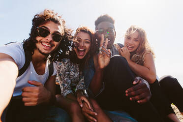 Aufgeregte junge Freunde, die ein Selfie im Freien machen. Verschiedene Gruppen von Männern und Frauen sitzen zusammen und machen ein Selbstporträt in ihrem Urlaub. - JLPSF00955