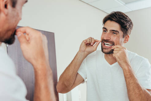 Mann schaut in den Badezimmerspiegel und benutzt Zahnseide, um seine Zähne zu reinigen. Reflexion des Mannes im Badezimmerspiegel beim Zähneputzen am Morgen. - JLPSF00957