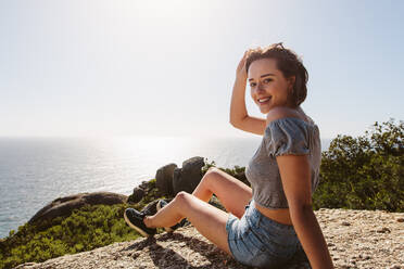 Lächelnde Frau sitzt auf dem Gipfel eines Berges und genießt die wunderbare Natur. Junge Frau auf Berggipfel mit schönem Meerblick. - JLPSF00964