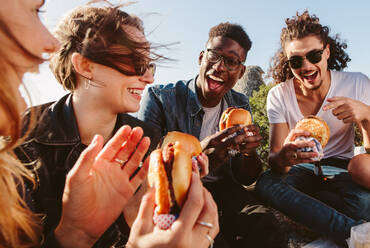 Group of friends sitting on mountain top eating burger. Excited young men and women enjoying and partying outdoors. - JLPSF00972