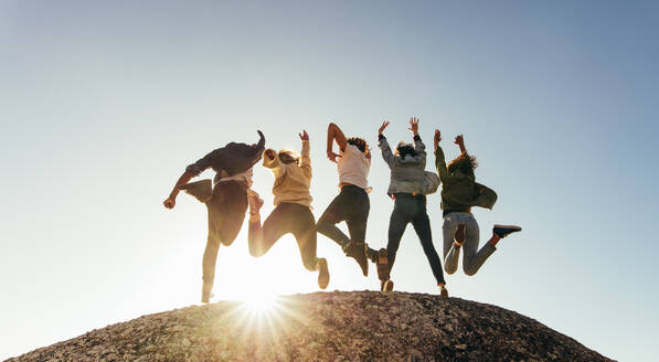 Rückansicht einer Gruppe glücklicher Freunde, die sich auf einem Berggipfel vergnügen. Männer und Frauen springen auf einem Berggipfel gegen den Sonnenuntergang. - JLPSF00977