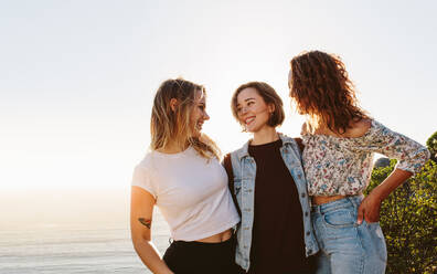 Three young women standing outdoors and looking at each other. Smiling female friends enjoying on summer vacation. - JLPSF00980