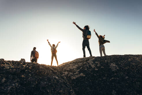 Rear view of friends having fun on mountain top and enjoying the view. Men and women standing on mountain top with their arms raised during sunset. - JLPSF00983