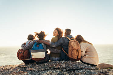 Eine Gruppe junger Männer und Frauen sitzt auf einem Berggipfel und bewundert den Sonnenuntergang. Eine junge Frau mit Freunden genießt einen Tag im Freien. - JLPSF00985