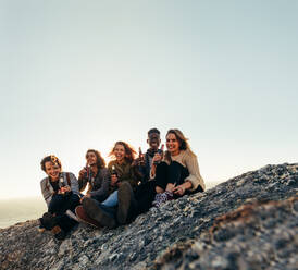 Multi-ethnic group of people with beers sitting on mountain top during sunset. Smiling young men and women enjoying beers and partying at sunset. - JLPSF00986