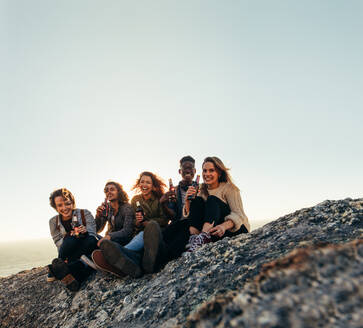 Multiethnische Gruppe von Menschen mit Bier auf einem Berggipfel während des Sonnenuntergangs. Lächelnde junge Männer und Frauen, die Bier genießen und bei Sonnenuntergang feiern. - JLPSF00986
