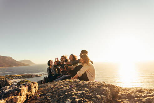 Freunde sitzen auf einem Berg und stoßen bei Sonnenuntergang mit einem Bier an. Eine Gruppe von Männern und Frauen feiert bei Sonnenuntergang im Freien. - JLPSF00991