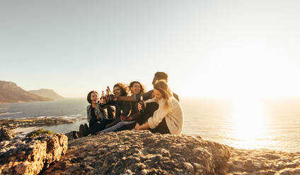 Group of friends having beers while sitting on mountain top during sunset. Multiracial men and women enjoying and partying outdoors. - JLPSF00992