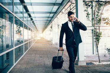 Gut aussehender junger Mann auf Geschäftsreise, der mit seinem Gepäck spazieren geht und am Flughafen mit seinem Handy telefoniert. Reisender Geschäftsmann beim Telefonieren. - JLPSF00995