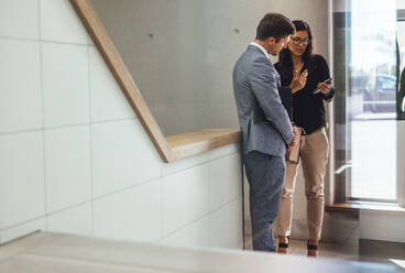 Zwei Geschäftskollegen stehen im Treppenhaus eines Büros und schauen auf ihr Handy. Männliche und weibliche Geschäftspartner diskutieren über ihre Arbeit und schauen auf ihr Smartphone. - JLPSF01028