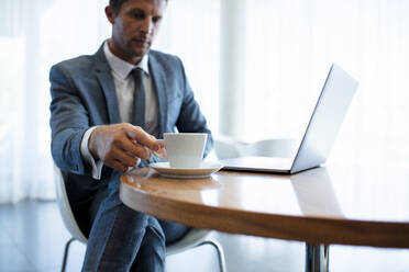 Geschäftsmann sitzt an einem Tisch mit Laptop und trinkt Kaffee, Fokus auf Mann, der eine Kaffeetasse in die Hand nimmt. - JLPSF01034