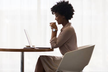 Afrikanische Geschäftsfrau lächelt, während sie einen Laptop benutzt und Kaffee im Büro trinkt. Seitenansicht eines Büroangestellten, der Kaffee trinkt, während er am Laptop arbeitet. - JLPSF01039