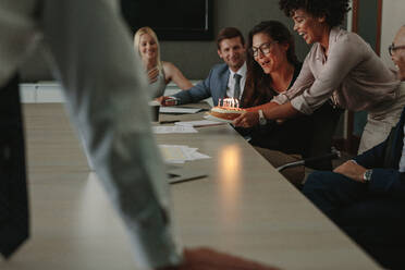 Coworkers congratulating female colleague on birthday and presenting cake with candles. Business people celebrating birthday of a female coworker in conference room. - JLPSF01057