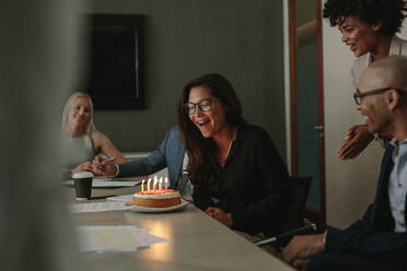 Surprise birthday celebration of a female associate during a staff meeting. Office workers surprising female colleague with cake on her birthday during meeting. - JLPSF01059