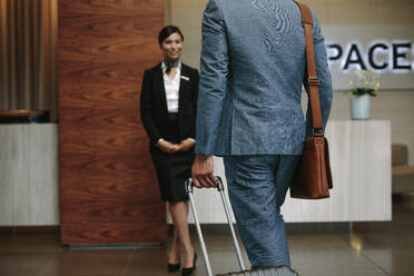 Geschäftsmann, der einen Koffer trägt und auf eine Empfangsdame zugeht, die auf die Begrüßung der Gäste wartet. Geschäftsreisender, der im Hotel für eine Konferenz ankommt. - JLPSF01062