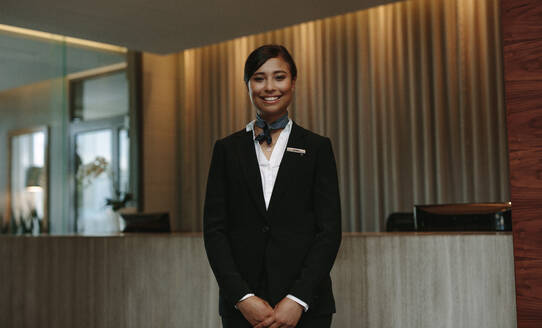 Junge lächelnde weibliche Rezeptionistin an der Hotelrezeption, die bereit ist, Gäste zu begrüßen. Weibliche Concierge schaut in die Kamera und lächelt. - JLPSF01066